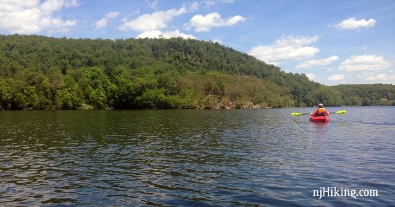 In the background, a tall hill rises behind a kayaker paddling Monksville Reservoir.