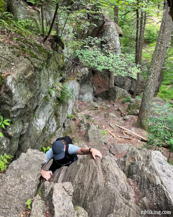 Hiker climbing up a rock scramble with the exit of the lemon squeezer behind him.