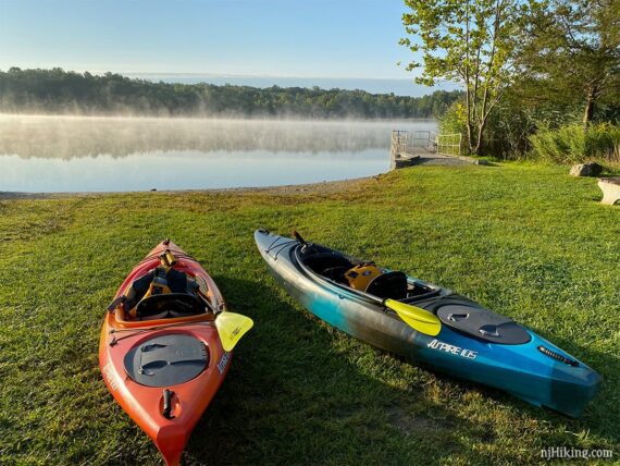 Two kayaks with paddles on grass at the edge of a lake.